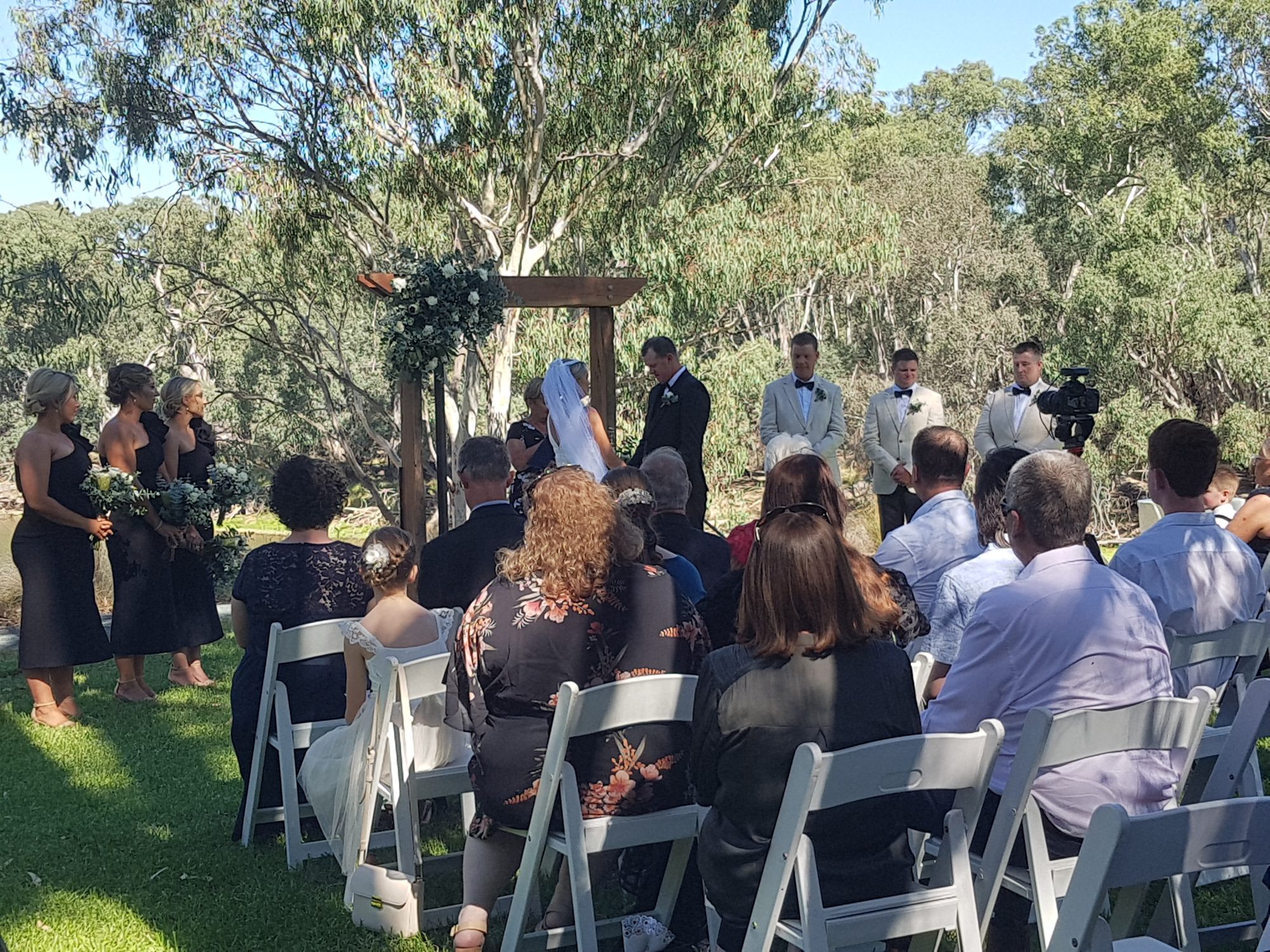Riverbank ceremony under the timber arch with guests seated on white chairs