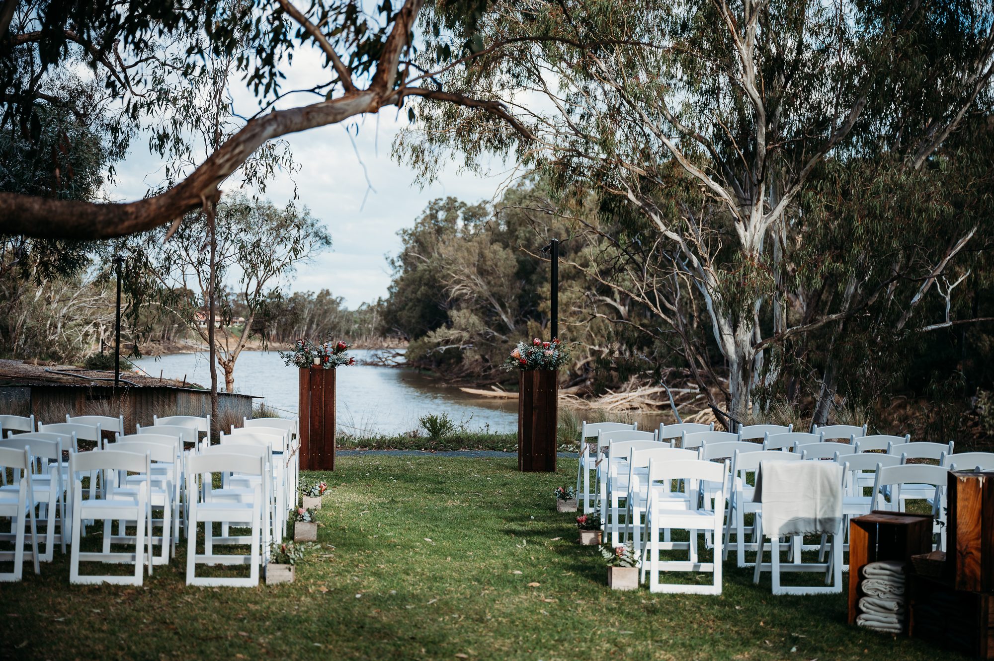 Riverbank ceremony aisle lined with white chairs and timber plinths, the river ahead